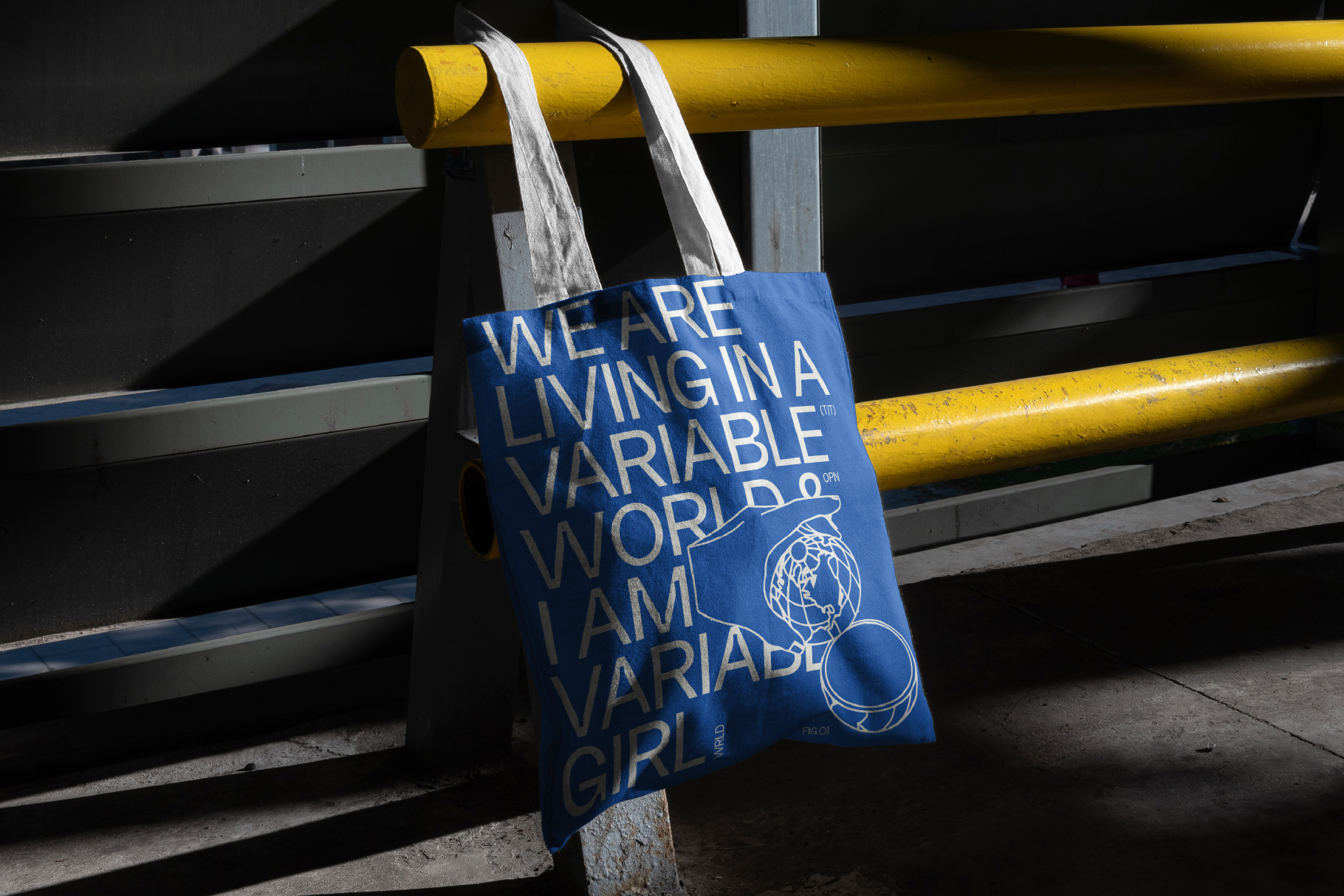 Blue tote bag with white text reading We Are Living In Variable World I Am Variable Girl hanging from yellow metal railing