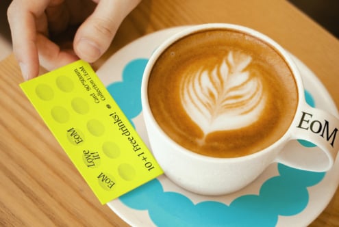 Hand holding a bright yellow loyalty card near a latte with latte art in a white cup on a wooden table ideal for mockup design graphic resources.
