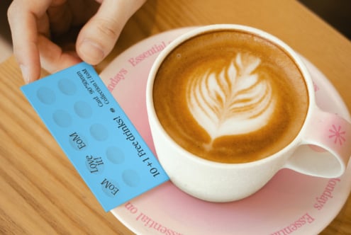 Hand holding loyalty card next to cappuccino in a cafe setting. Ideal for coffee shop graphics, branding mockups, and template designs for designers.