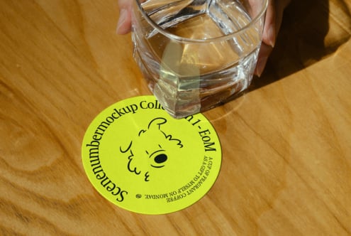 A hand holding a glass on a wooden table with a round yellow coaster mockup featuring playful typography and a simple illustration for graphic designers.