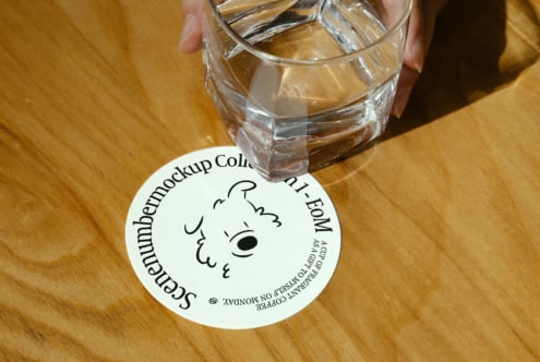 Mockup of a hand holding a glass on a wooden table, featuring a round paper coaster with text; ideal for product presentation and branding design.