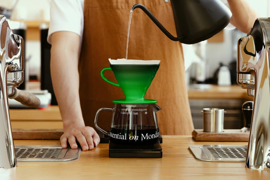 Bright green ceramic dripper with white filter pouring into black glass carafe on wood desk