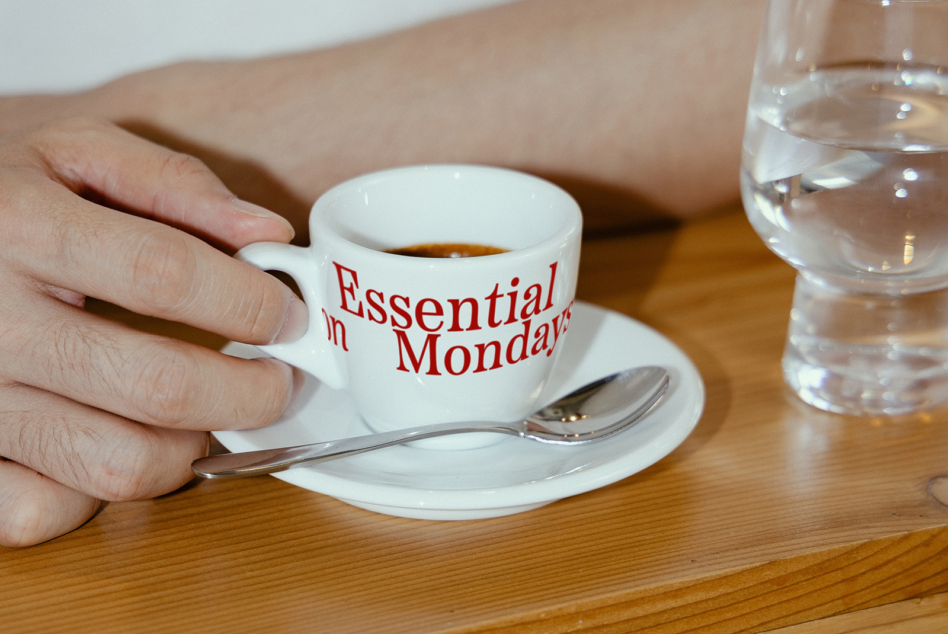 Red text on white espresso cup reading Essential on Mondays, held in hand