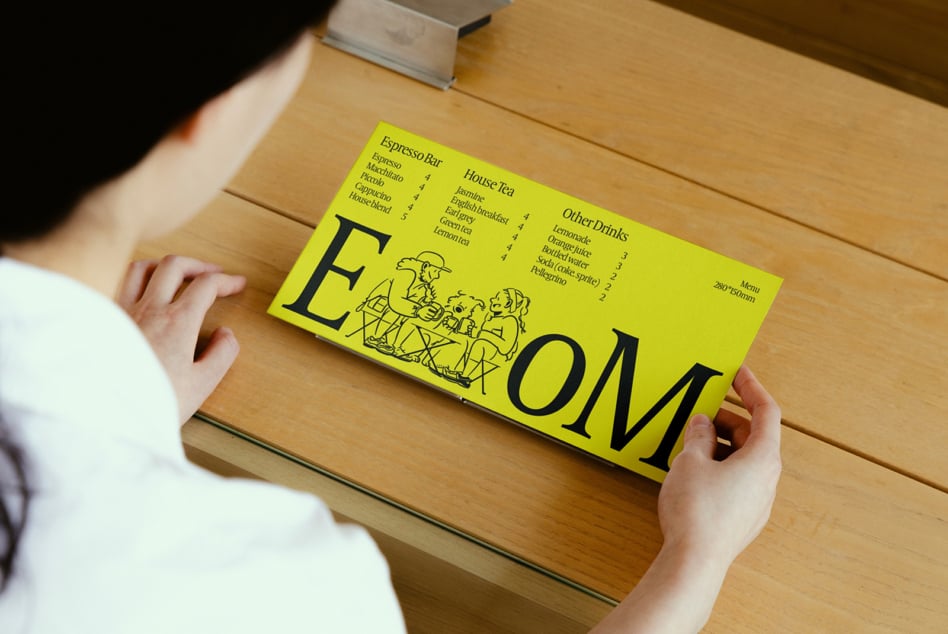 Bright yellow cafe menu mockup with hand-drawn people illustration, held in hands over wooden table