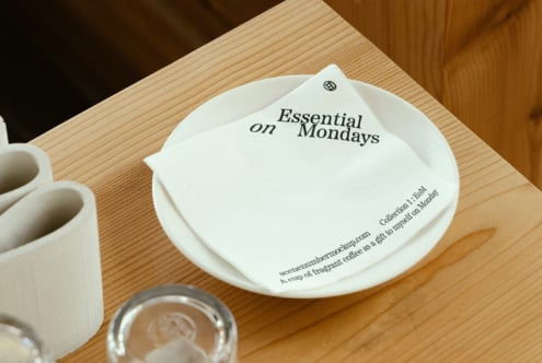 Mockup image featuring a white napkin printed with Essential on Mondays on a wooden table for graphic design projects and branding presentations.