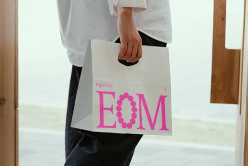 Person holding a white shopping bag with pink letters EOM design. Perfect for mockups or graphics. Ideal for branding and marketing projects.