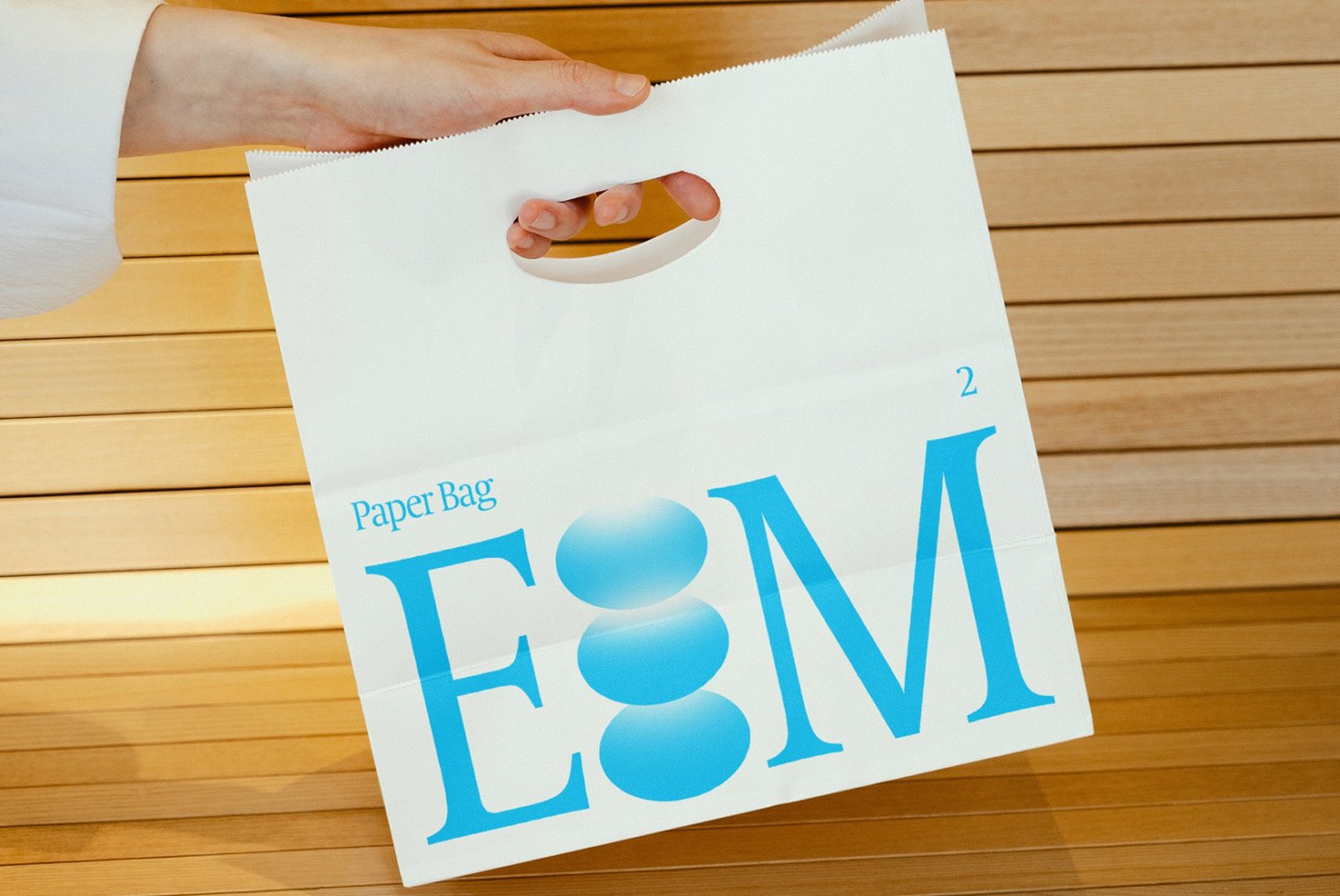 Mockup of a white paper bag with large blue typography design held by a hand against a wooden background. Perfect for branding and packaging design.