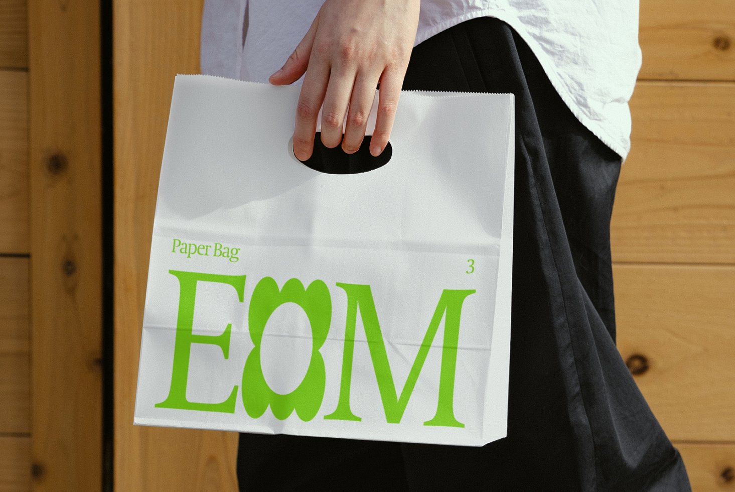 Person holding a white paper bag with bold green typography EOM. Ideal for mockups, branding, shopping design assets, packaging templates, eco-friendly design.