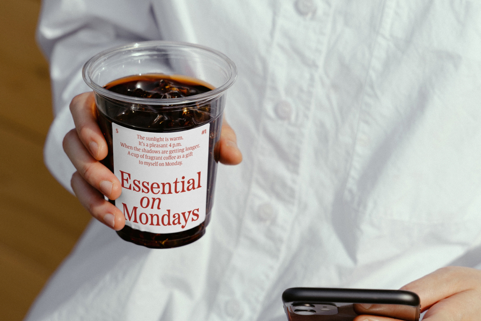 Hand holding clear plastic cup with red label Essential on Mondays over iced coffee