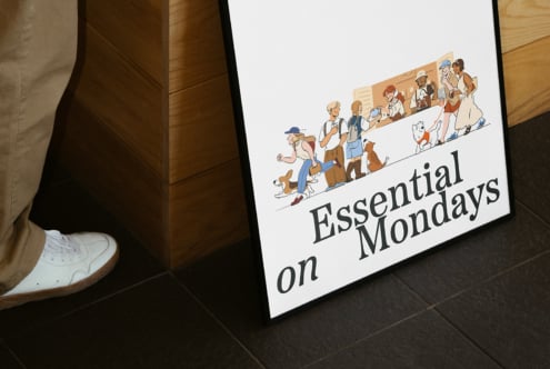 Framed poster with cartoon characters and Essential on Mondays text; leaning against a wooden surface; ideal for graphic designers and mockup creators.