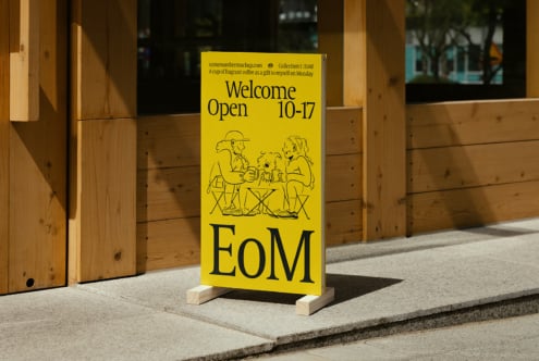 Bright yellow sign mockup with black text and illustrations displayed outside a wooden building. Ideal for showcasing custom fonts or retail signage design.