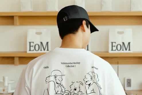 Man in black cap wearing white T-shirt with cartoon design stands in front of shelves. Ideal for mockup display, apparel showcase, graphic design.