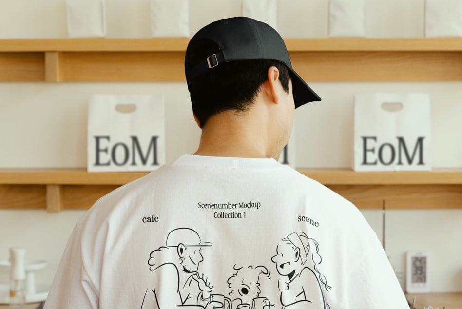 Man in black cap wearing white T-shirt with cartoon design stands in front of shelves. Ideal for mockup display, apparel showcase, graphic design.