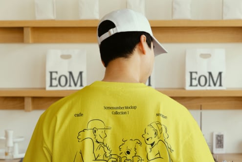 Man in yellow graphic tee and white cap from behind in a cafe setting. Design mockup with playful characters. Ideal for trendy clothing mockups.