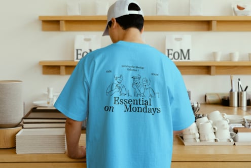 Man wearing blue t-shirt with printed graphic and Essential on Mondays text in a modern cafe setting. Perfect for mockups or graphic design templates.