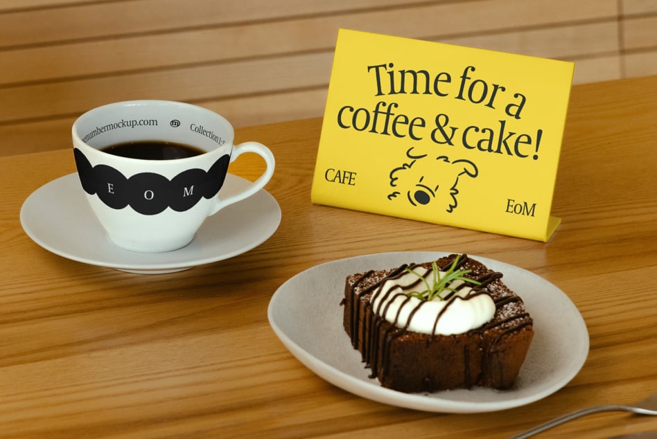 Café setup with white cup featuring mustache design, yellow sign, chocolate dessert on wooden table