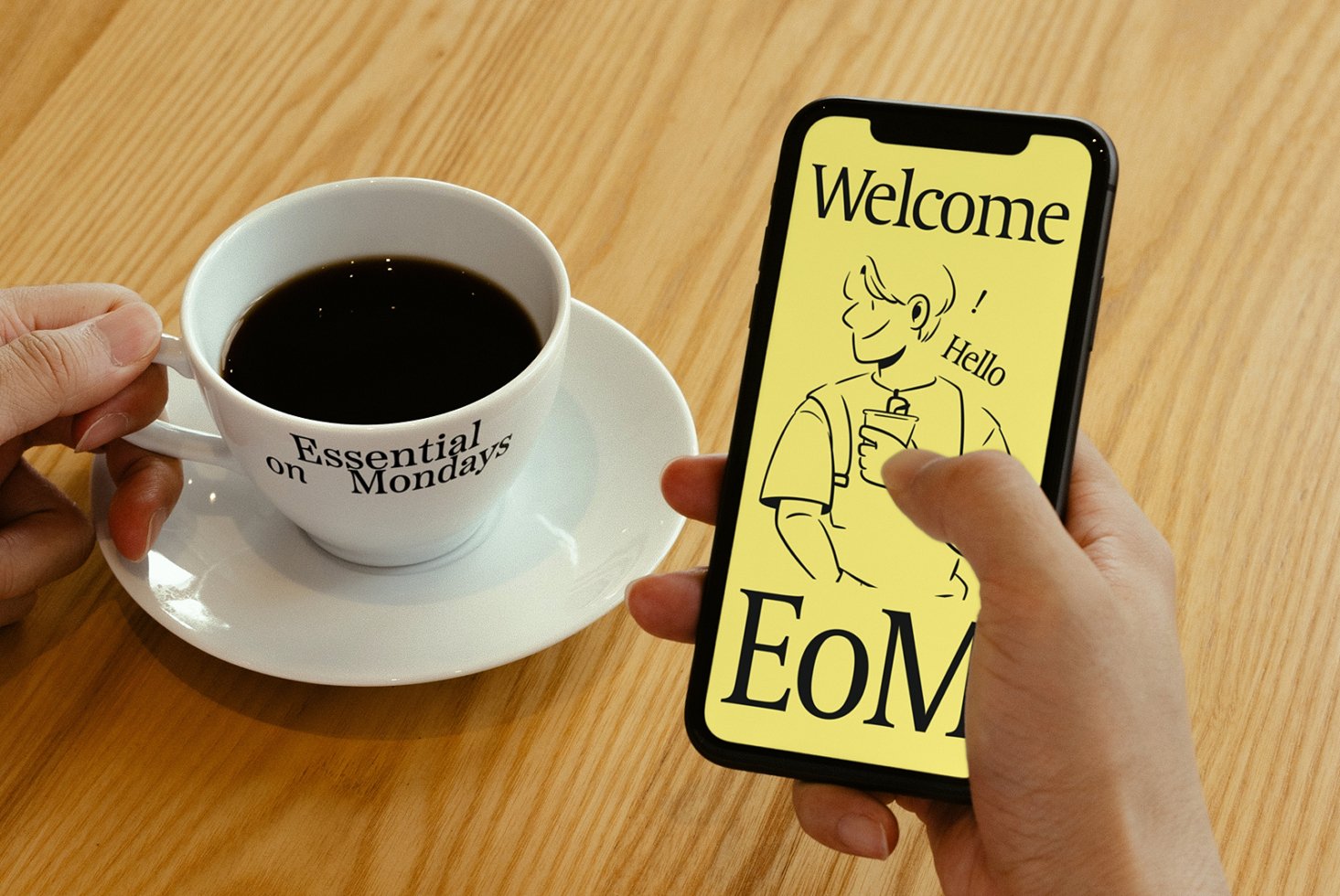 Person holding a smartphone with a welcome design on screen, beside a coffee cup labeled Essential on Mondays. Ideal for mockups and templates.