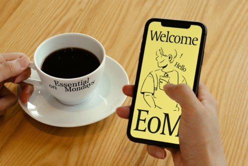 Person holding a smartphone with a welcome design on screen, beside a coffee cup labeled Essential on Mondays. Ideal for mockups and templates.