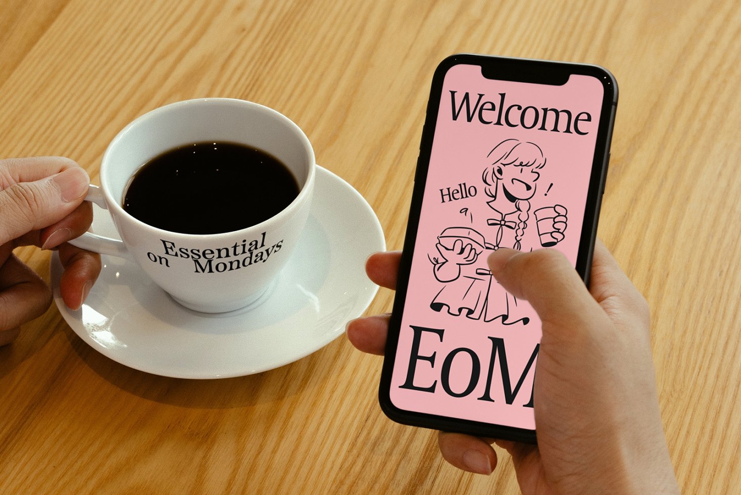 Smartphone displaying pink welcome screen with cartoon figure alongside coffee cup reading Essential on Mondays over wooden table. Mockup design template.