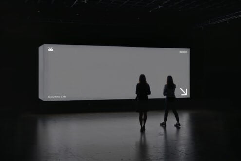 Two silhouetted figures stand before a large, illuminated screen displaying text Colortime Lab in a dark room. Keywords: mockup, minimalist, display.