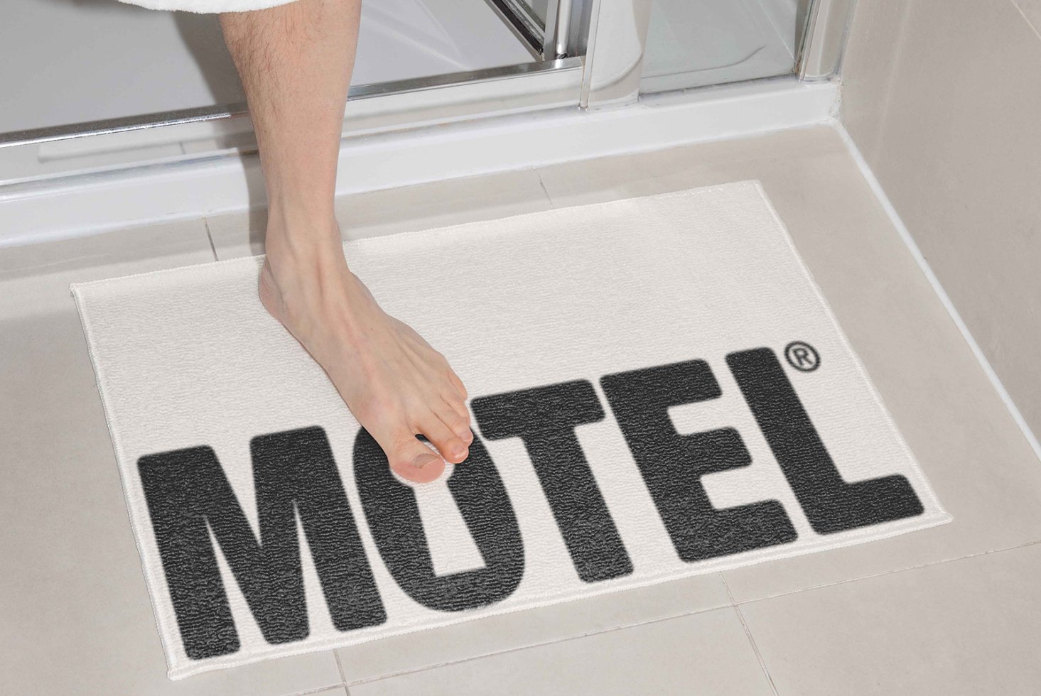 Bath mat mockup with foot stepping out of shower onto fabric labeled Motel. Ideal for digital designers seeking realistic textured designs for templates.