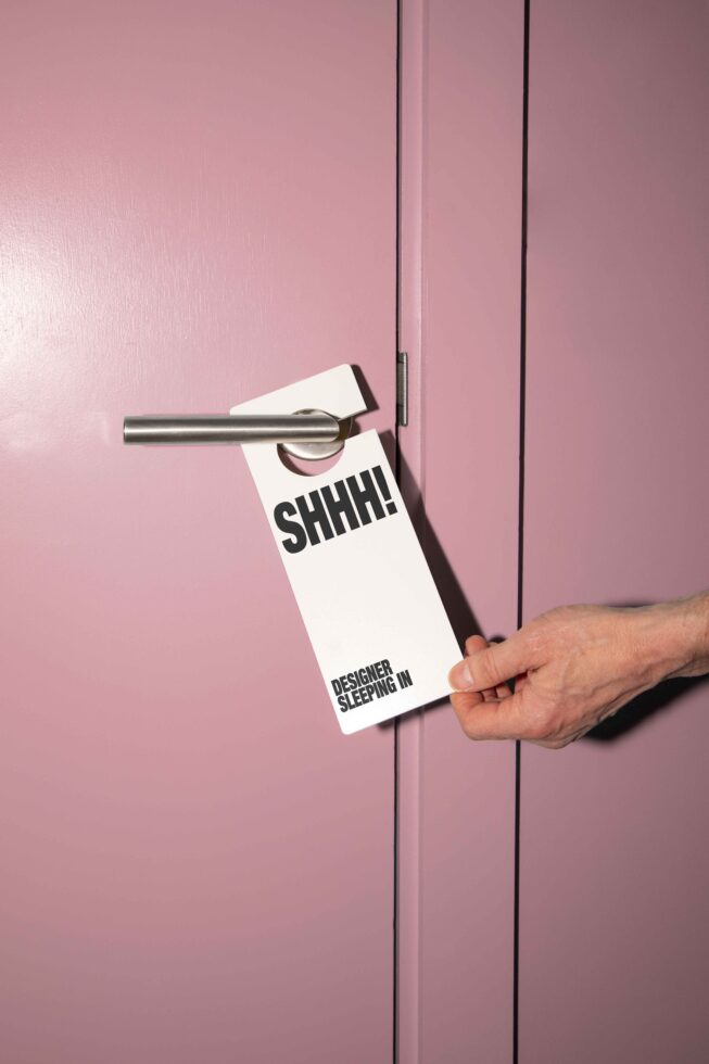 Pink door with a silver handle and a person placing a door hanger reading Shhh Designer Sleeping In. Ideal for mockups, graphics, templates, design assets.