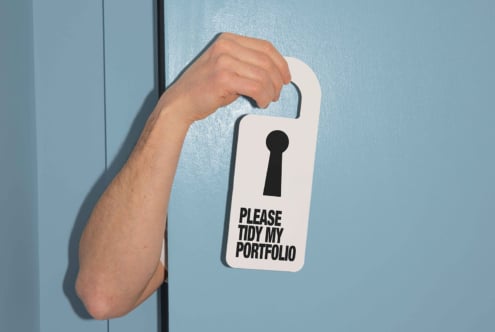 Hand holding a door hanger reading please tidy my portfolio against a blue door. Perfect for design mockups and creative templates. Ideal for designers.