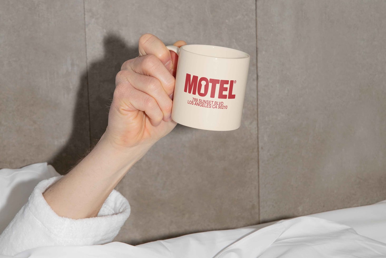 Hand holding cream mug with motel address text against gray wall