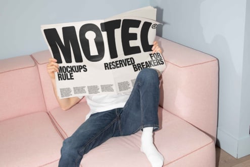 Person reading newspaper with bold MOTEL headline on pink couch blue wall. Perfect for mockups marketing designers typography modern interior.