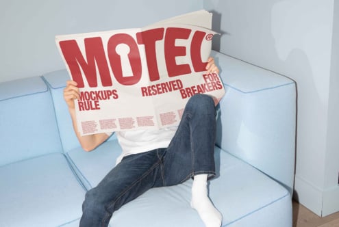 Person on blue sofa reading large red-lettered newspaper. Mockups rule text visible. Ideal image for graphic designers. Creative workspace concept.