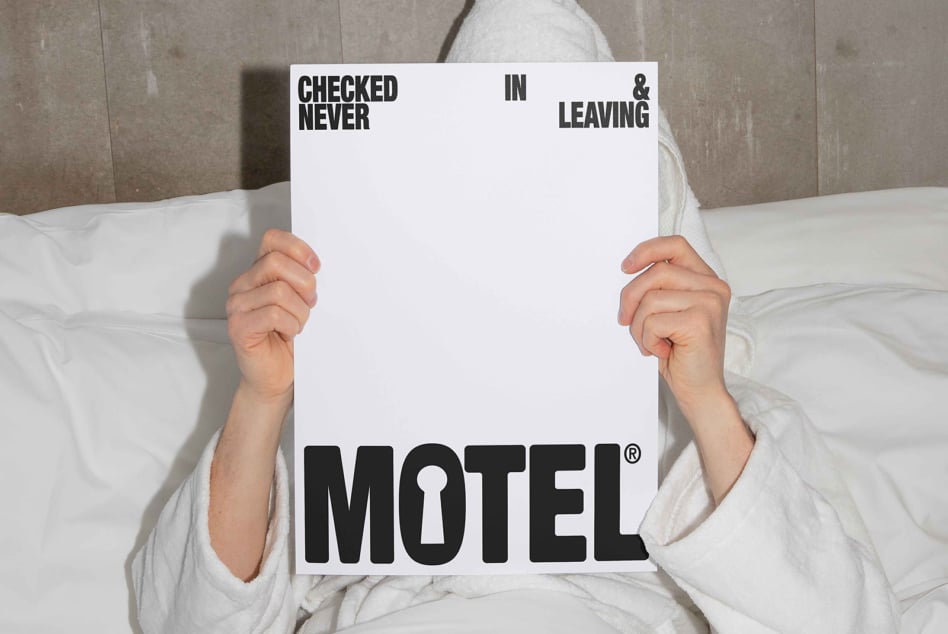 Person in bed holding black Checked In Never Leaving Motel sign