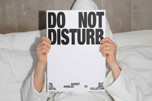 Person in white robe holding a DO NOT DISTURB sign while laying in bed. Suitable for graphics or mockups for designers focusing on typography.