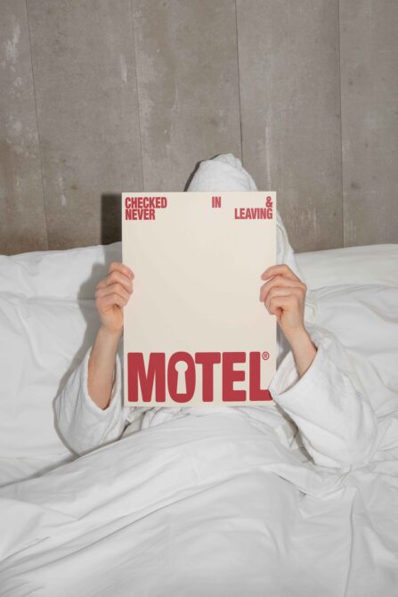 Person in white bedding holding red Checked In Never Leaving Motel sign