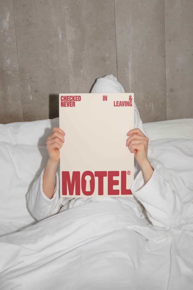 A person in a robe holds a poster in bed displaying Motel with bold red text. Suitable for mockups or graphic design templates in hospitality themes.