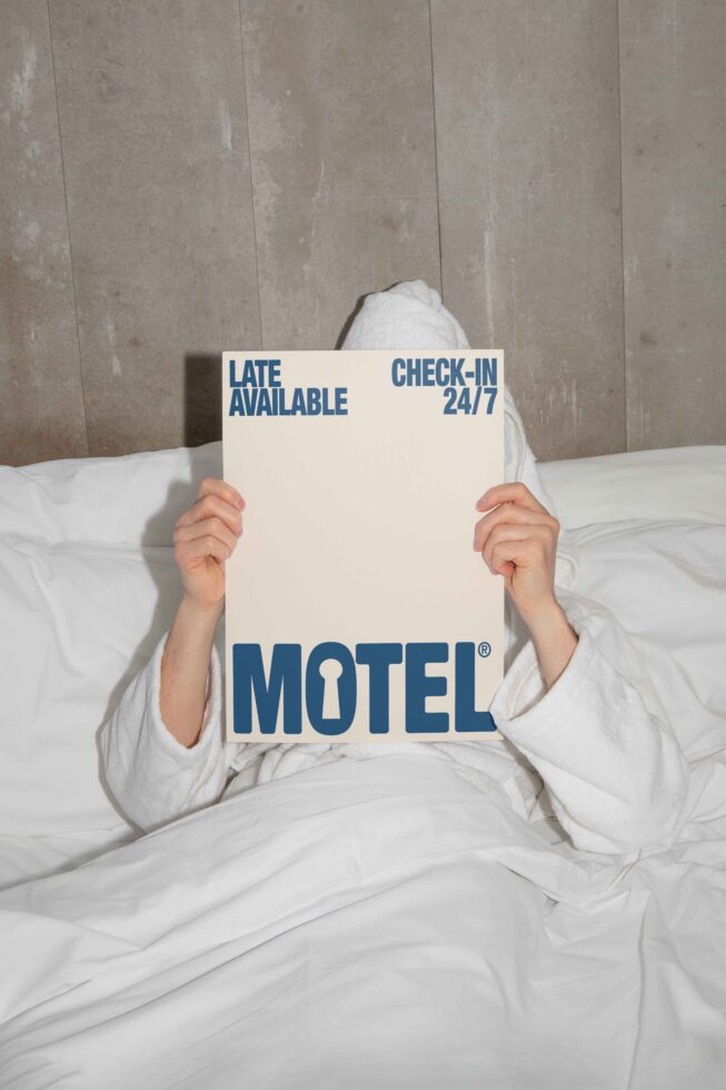 Person in a white robe on a bed holding a motel sign with bold blue text reading Late Available Check-in 24/7 ideal for hotel advertising mockups.