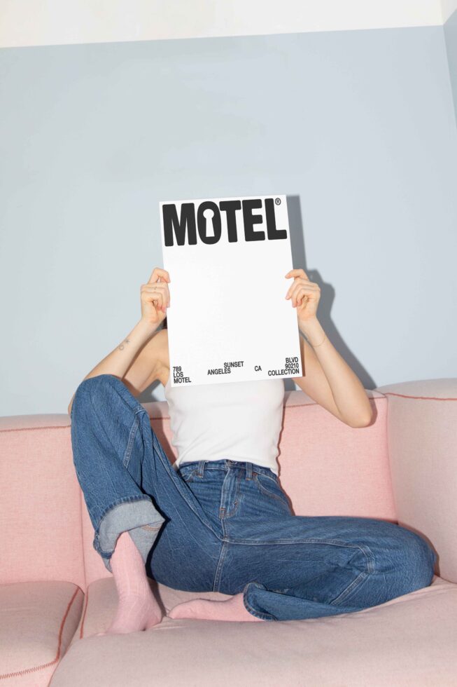 Person holding Motel poster on a pink sofa in denim jeans and white top. Modern graphic design inspiration for mockups and creative projects.