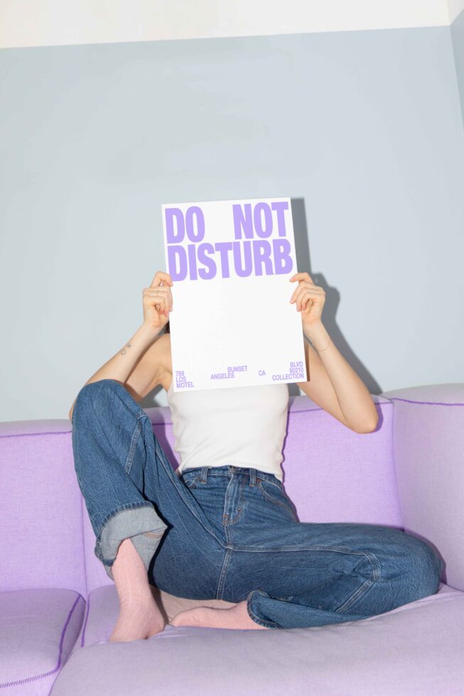 Person sitting on a purple couch holding a Do Not Disturb sign covering face; denim jeans, casual attire; perfect for mockup design; indoor setting.