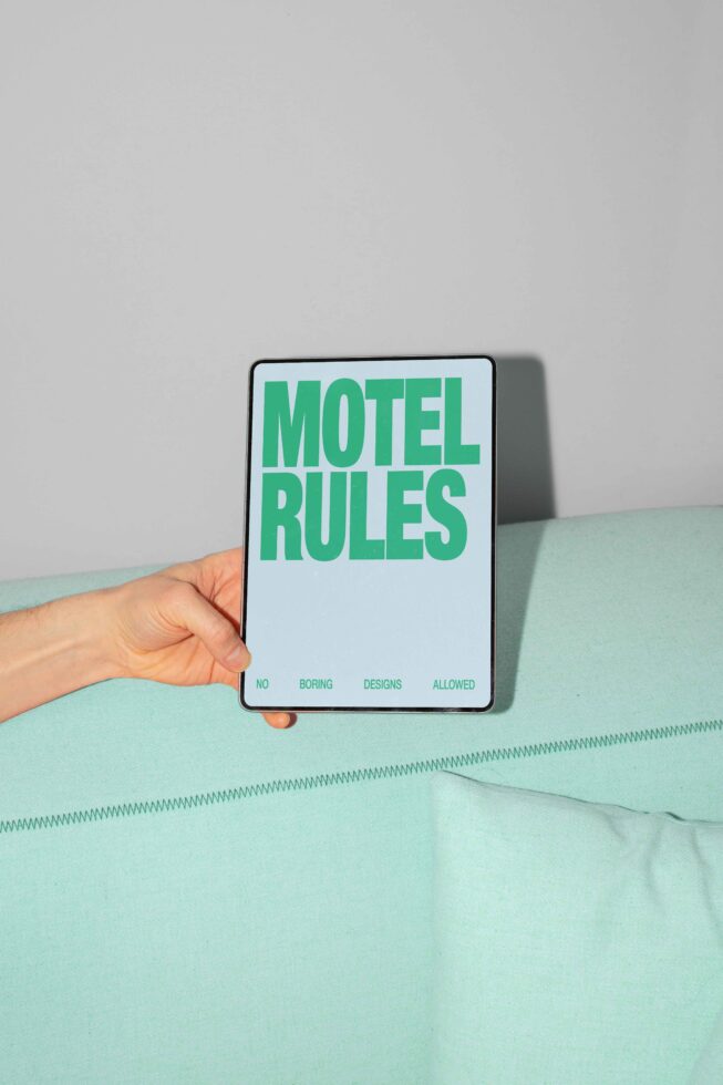 Hand holding a tablet with Motel Rules text in bold green on a pastel aqua fabric background. Perfect for designers seeking quirky mockups or templates.