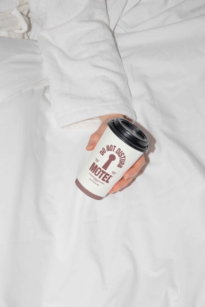 Hand in white robe holding coffee cup with Do Not Disturb Motel design on white bed background. Ideal for mockup presentations, graphic design resources.