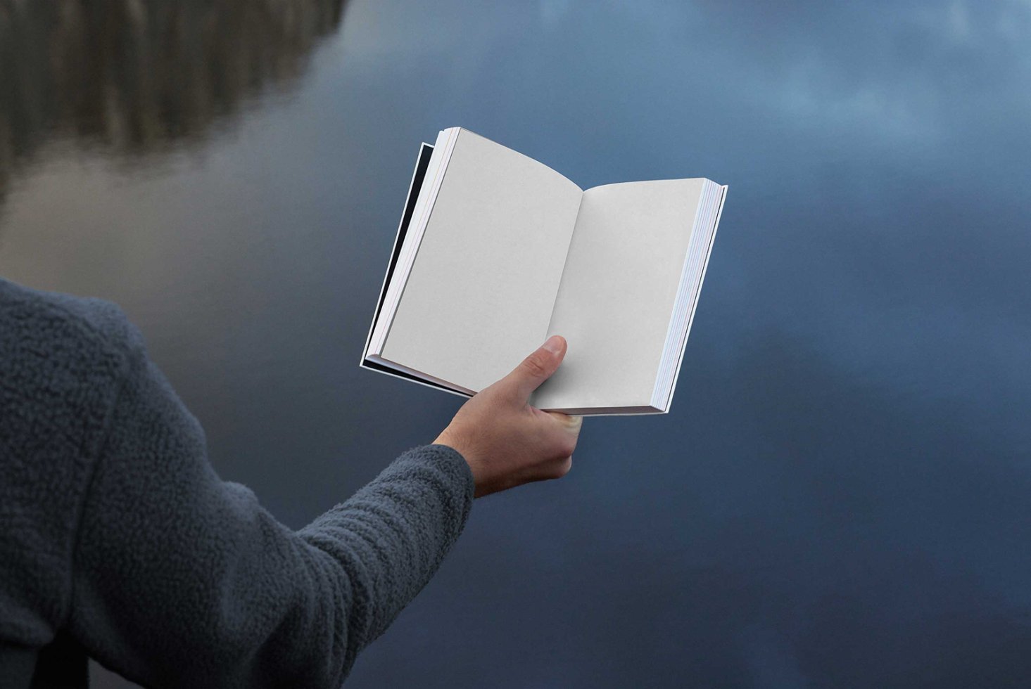 Open book mockup held by a person in front of blurred water background. Perfect for showcasing cover designs. Ideal for designers and graphic projects.