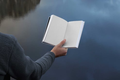 Open book mockup held by a person in front of blurred water background. Perfect for showcasing cover designs. Ideal for designers and graphic projects.