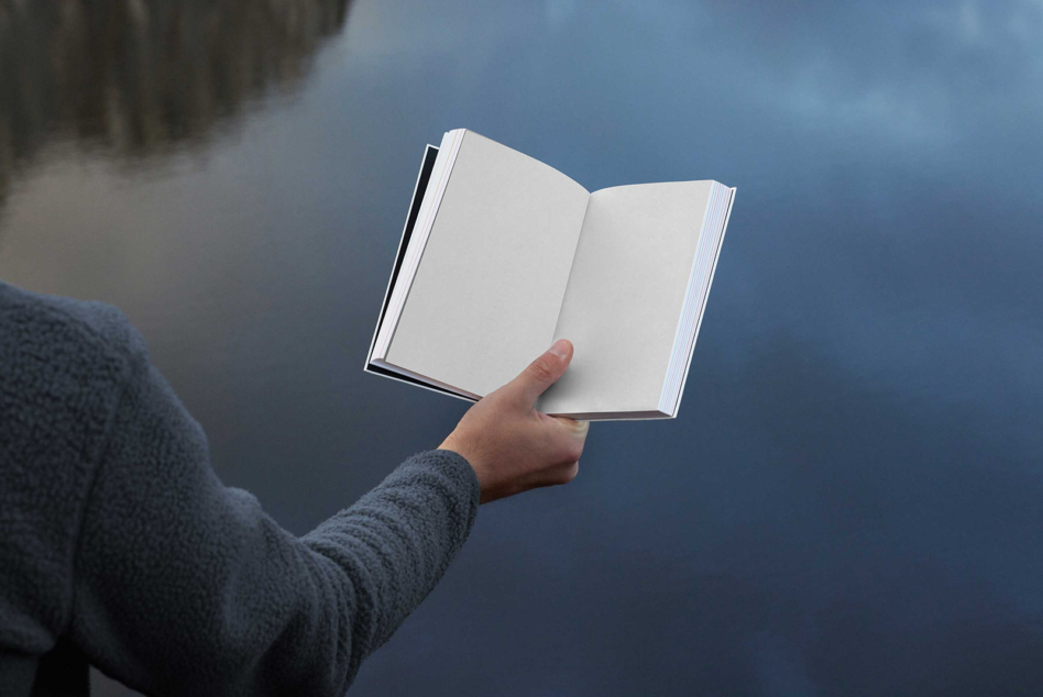 Open hardcover book with blank pages held by hand against blue water backdrop