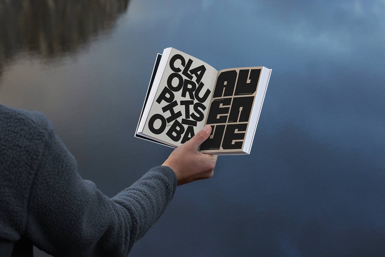 Book mockup with bold typography design held by a person over a reflective water surface background ideal for graphic designers and typographers.