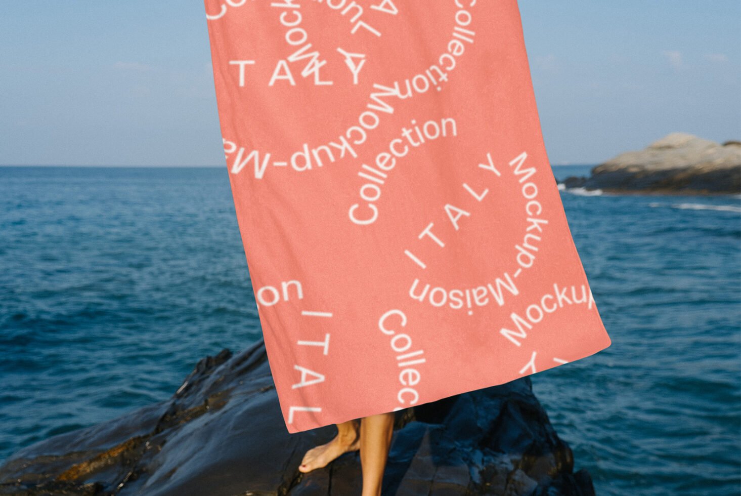 Beach towel mockup featuring a coral color with white text design Italy Collection near the ocean rocks ideal for showcasing textile graphics