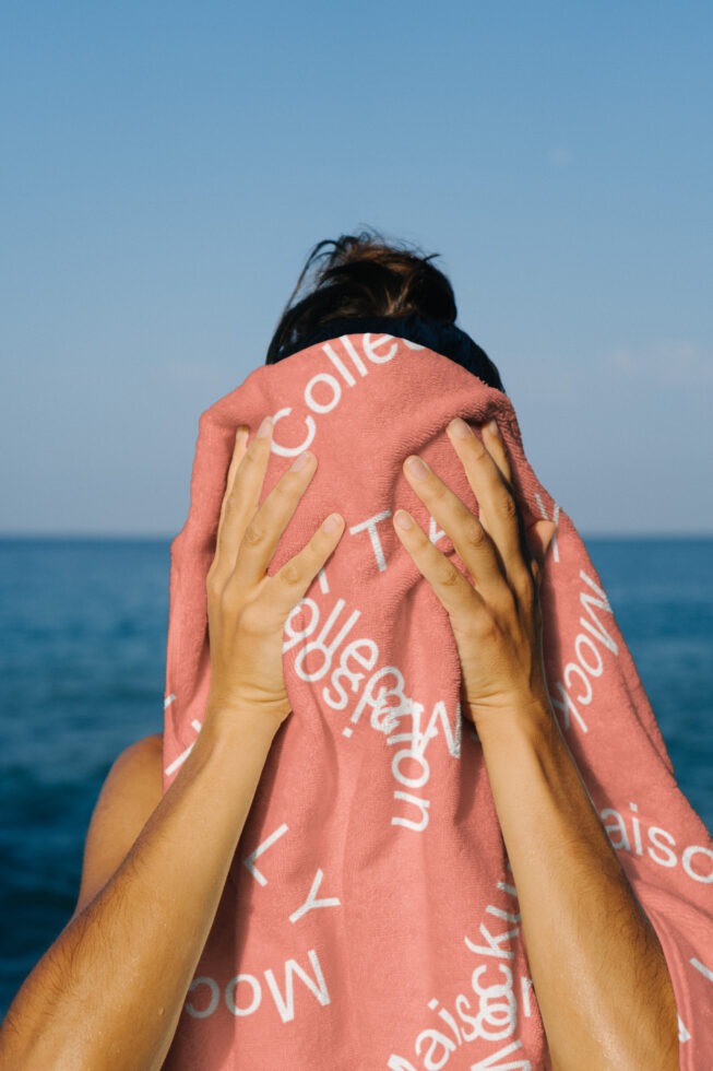 Person holding pink towel with white letters near ocean blank face clothing mockup summer design template blue sky keywords for designers.