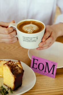Hands holding magenta business card next to coffee cup with pastry on wooden table