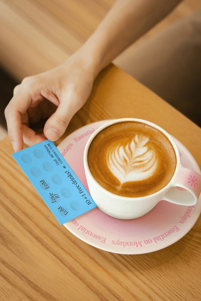 Latte art coffee cup on a saucer with hand holding blue loyalty card over wooden table, perfect for branding mockups and design templates