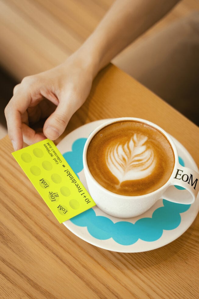 Hand holds loyalty card next to coffee cup with latte art on wooden table. Perfect for mockups, branding, cafe designs. Eye-catching design element.