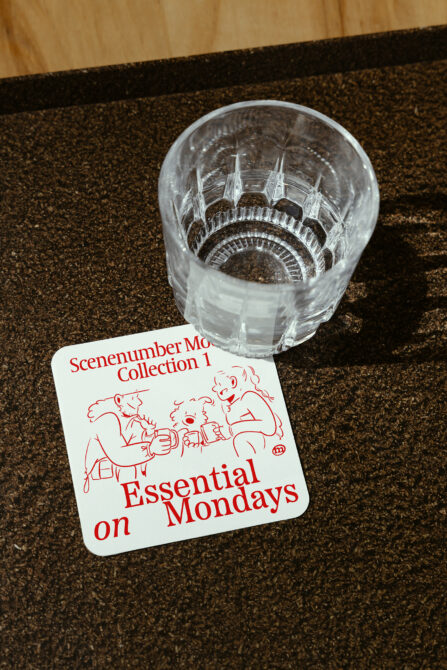 Transparent drinking glass on cork coaster with red text label featuring cartoon figures