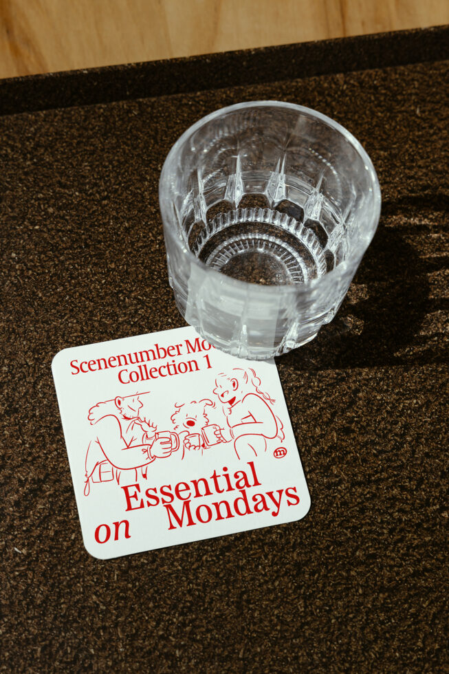 Glass tumbler on dark textured surface next to a white coaster with red text and illustrations. Perfect for mockup or graphic design enthusiasts.